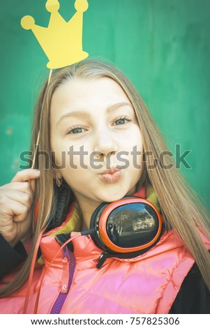 
Portrait of a teenage girl with headphones and a paper crown and making a face, against a green wall background