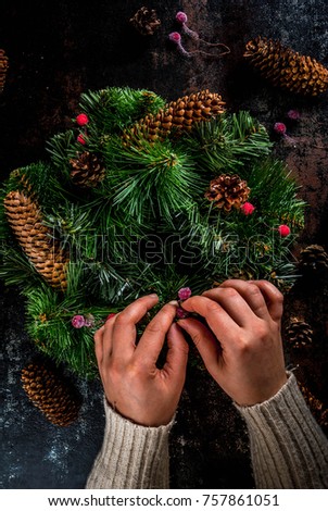 Preparation for xmas holidays. Woman decorating christmas green wreath with pine cones and red winter berries, on dark rusty  background, top view copy space, female hands in picture
