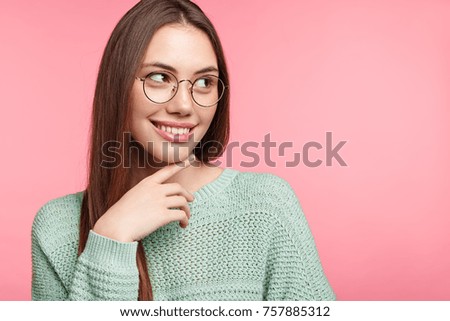 Lovely young Asian female has happy thoughtful expression, looks aside, notices handsome guy before, feels shy, poses against pink background with copy space for your text or advertising content