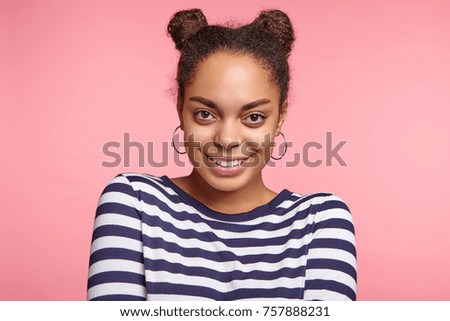 Happy African American young woman with hair knots, wears round earrings, looks directly into camera, to meet international friends, isolated over pink background. People and happiness concept