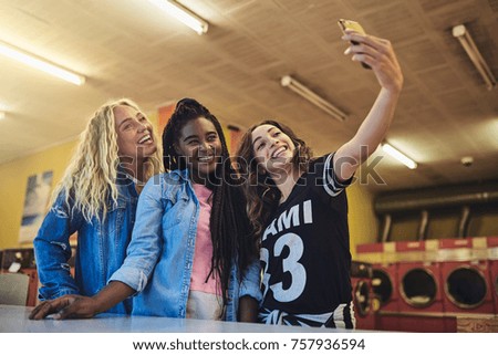 Smiling young female friends standing in a laundromat taking a selfie while doing laundry together