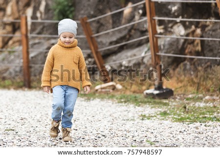 Little girl with grey hat, orange sweater, dirty jeans and boots walks on puddle on the background of fence. Time autumn or winter.