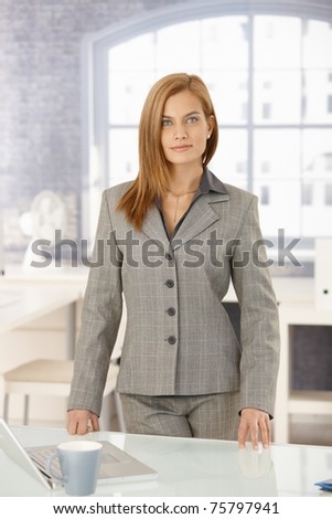 Portrait of confident businesswoman in office, standing at desk, smiling at camera.?