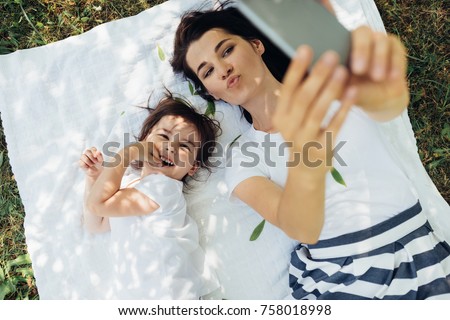 Top view of happy young beautiful mother blowing kiss and taking self portrait with cute daughter smiling during picnic lying on grass and white blanket. Motherhood, technology and childhood concept.