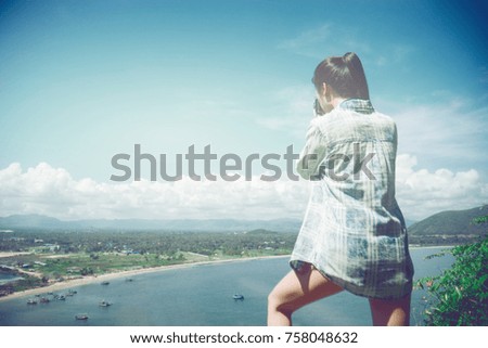 portrait of asian woman photographer shooting photo of view high view from a cliff mountain. blue sea and blue sky with white cloud. filtered image. selective focus. light effect added. travel concept