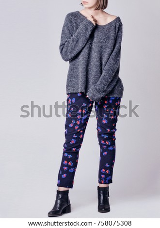Woman wearing stylish casual outfit with black patterned floral cigarette trousers, big gray oversized jumper and black ankle boots isolated on grey background. Copy space
