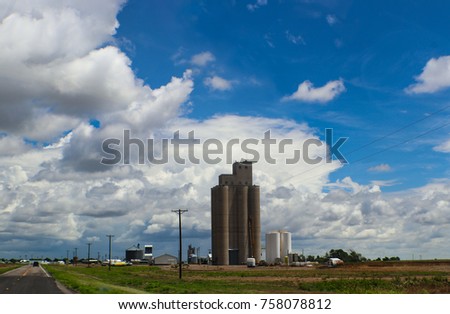 Tall grain elevators in the midwestern United States sorrounded by trucks and equipment and powerlines beside a highway all under a huge blue sky with fluffy clouds