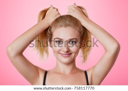 Photo of funny joyful female model makes two pony tails from hair, stands in front of mirror, thinks what hairdo to make as going on party with friends. Preparation for special event. Having fun