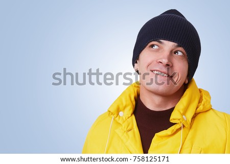 Stylish hipster guy wears fashionable yellow anorak and black hat, looks pensively aside, dreams about something pleasant, isolated over blue background. Cheerful young man poses in studio alone