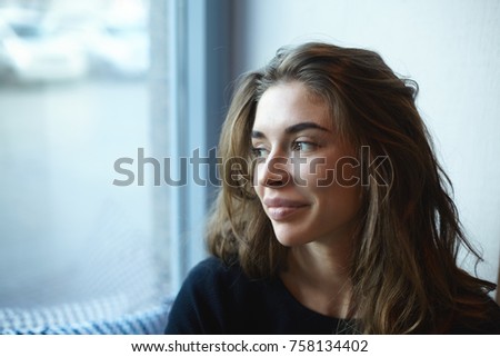 Sideways view of charming pretty girl with healthy skin and voluminous hairstyle enjoying good day indoors at cafe, looking through window and smiling joyfully as she sees her friend outside