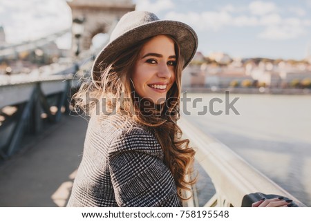 Outdoor photo of romantic european woman with curly hairstyle spending time outdoor, exploring european city. Graceful young lady in gray coat and hat enjoying views on embankment. Royalty-Free Stock Photo #758195548