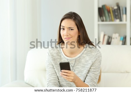 Front view portrait of a suspicious woman holding phone looking at you sitting on a sofa in the living room of a house interior Royalty-Free Stock Photo #758211877