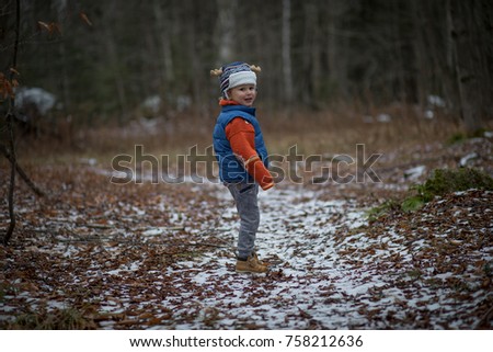 A little boy is hiking in the woods on a winter day on a snowy trail. He has a moose hat and a blue jacket vest with an orange sweater.  There are fallen leaves in the forest where he is hiking. 