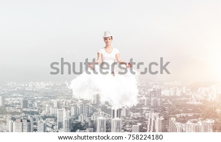 Woman in white clothing keeping eyes closed and looking concentrated while meditating on cloud in the air with cityscape view on background.