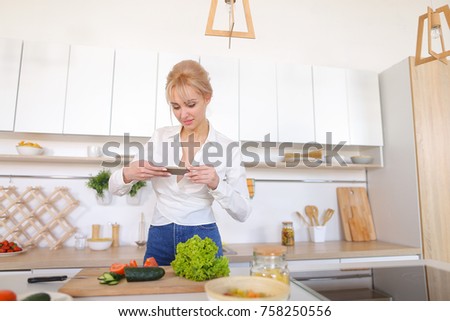 Charming woman using mobile gadget records video of cooking process and photographs composition of tomatoes and cucumbers with salad leaves on cutting board, vases with apples and bananas for female