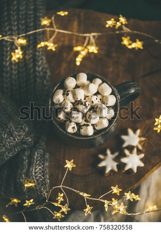 Christmas or New Year winter hot chocolate with marshmallows and gingerbread cookies over wooden board served with holiday light garland, blanket and grey sweater, selective focus, top view