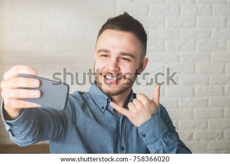 Portrait of attractive young man with call me gesture, taking selfie with smartphone in blurry office. Success and hipster concept 
