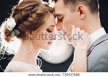 Portrait of beautiful young couple young gorgeous bride and handsome groom at rounded light background. Sweet, beloved sweathearts leaning by nose each other, embracing and touching by faces. Wedding. Royalty-Free Stock Photo #758437888