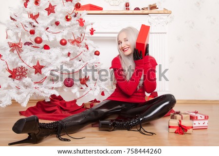 Beautiful Fashionable Blonde Woman Wearing Black Latex Rubber Catsuit, Fetish High Heels Boots and Red Lond Sweater Posing Near The White Christmas Tree and FirePlace With New Year Boxes Of Presents