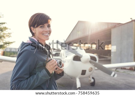 Smiling young female photographer and tourist at the airport, she is holding a digital camera, propeller plane and hangar on the background