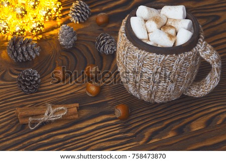 Cup of hot chocolate with marshmallows, nuts and cinnamon on a vintage wooden surface with christmas lights on background, selective focus