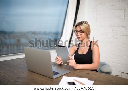 Young woman commercial real estate analyst reading e-mail on touch pad, sitting at the wooden table with laptop computer. Female secretary big successful company using digital tablet and net-book 