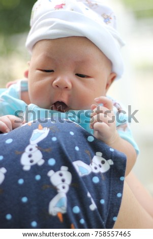 A picture portrait of an Asian Baby boy. He was carried by his mom and walk to the park in the morning under sunlight. Baby is wearing blue shirt and white hat. He open his mount like to say something