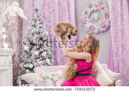 Beautiful young girl with long wavy hair in a pink dress on a background of a Christmas tree holding a rabbit in her arms. Christmas and New Year in the image.