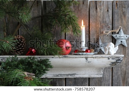 Christmas composition with burning candle in vintage enamel candlestick , red glass decorations, cone, porcelain angel on metal stars on aged wooden background, fresh brunches of pine on old shelf