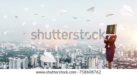Cropped image of business woman in suit with monitor instead of head keeping arms crossed while standing outdoors among flying paper planes with cityscape view on background.