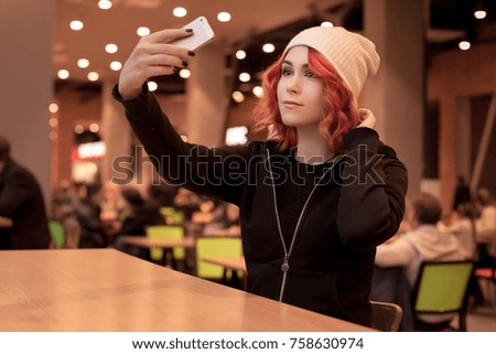 Beautiful young girl, female student with coffee sitting on food court in a shopping center. He makes a selfie, a photo of himself and listens to music on headphones. Orange hair.