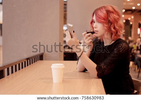 Beautiful young girl, female student with coffee sitting on food court in a shopping centerYou corrects make-up, paints lipstick, looking in reflection in the phone. Orange hair. Narcissism.