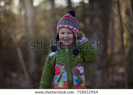 A little boy is in the woods with a winter holiday hat and an Embroidered Peruvian sweater. He is smiling and happy on a fall or winter day. Trees and sunlight are in the background of the photo.  