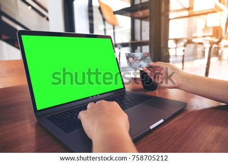 Mockup image of a hand holding credit cards while using and typing on laptop with blank green screen and coffee cup on wooden table in modern loft cafe