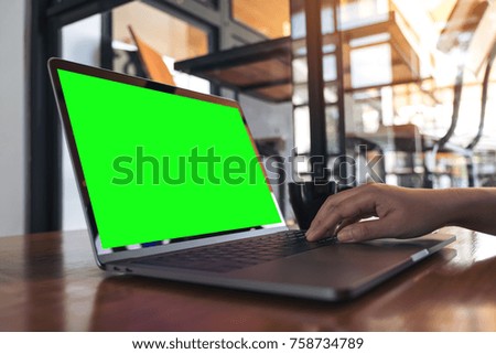 Mockup image of a hand using and typing on laptop with blank green screen and coffee cup on wooden table in cafe