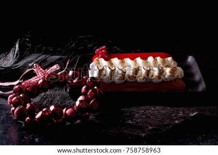Home made eclairs decorated with white meringue and red icing on the square plate with red decoration on the black background. Dark Rustic style.