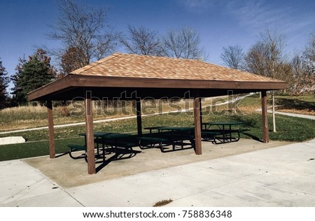Horizontal full color photo of picnic area with tables and roof canopy on concrete patio in the park with trees, grass and walking path in the background