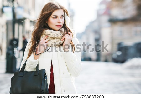 Outdoor portrait of young beautiful fashionable woman wearing stylish white winter puffer coat, scarf, holding leather tote bag. Model walking in street. Female fashion concept. Copy, empty space