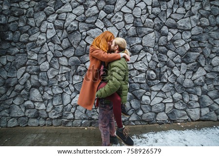 Happy people relationship concept. Young stylish couple embrace at grey wall urban background. Man lift up his redhead girlfriend.
