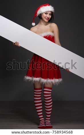 beautiful young woman in a red skirt and Santa Claus hat holds a white poster with a copyspace place for the text for your advertisement