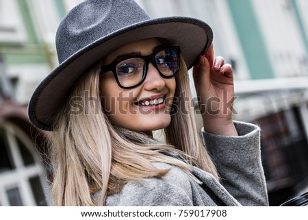 Cheerful elegant woman. Portrait of beautiful stylish woman in coat adjusting hat on head and smiling while standing outdoors. Fashionable hipster girl walking in the city. Urban fashion style.