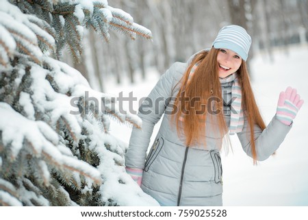 Cheerful girl looks out from behind trees and waving his hand, looking at the camera.
