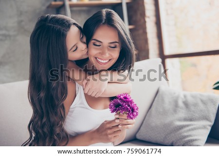 Concept of women's day! Amazed cheerful joyful charming mum is holding nice flowers and is receiving a kiss from her little cute daughter with long curly hair