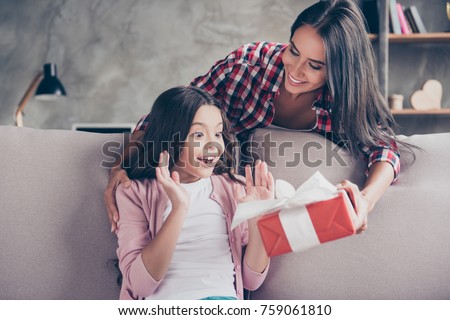 Dreams come true on birthday! Here you are a present! Young charming mother in casual clothes is giving a red gift box to her surprised and cheerful little princess