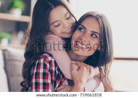 Playful small girl with long dark hair is hugging her mum's neck and looking at her with a smile, they have perfect weekends at home