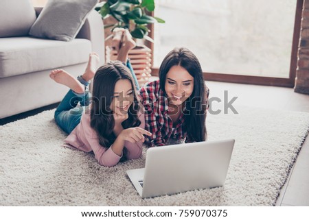 Cheerful toothy smiling happy mother and her little curious daughter are lying on a floor at home and usinf a laptop for watching cartoons, a girl is indicating on the monitor at the weekend