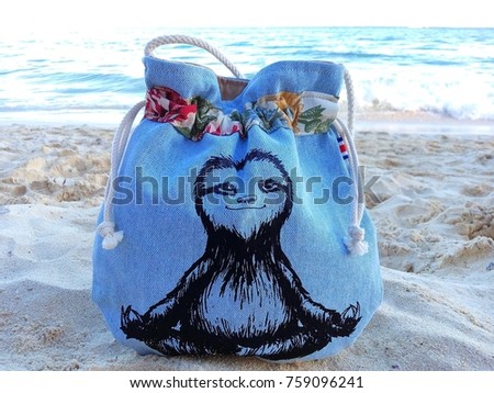 Close-up of traditional handmade Costa Rican blue jeans bag with funny picture of smiling meditating sloth on white sandy beach with Caribbean Sea in background – concept of souvenir or exotic holiday