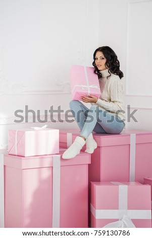 Young cute teenage girl in knitted white sweater and socks. She holds gift in her hand and sits on large pink gift. Holidays. Happy New Year. Merry Christmas. Valentine's Day. Birthday.