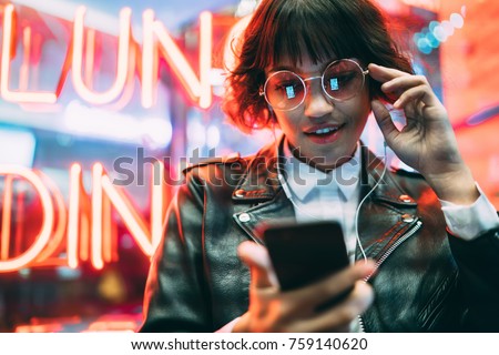 Attractive brunette woman in spectacles searching songs in network for downloading while standing near night city lights,positive hipster girl watching online video on phone using earphones outdoors Royalty-Free Stock Photo #759140620