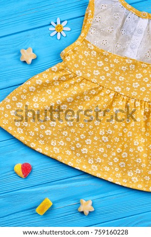 Baby-girl yellow sundress. Cotton summer dress in small white flowers for little girls on blue wooden background. Nice decoration from rubber candies near kids clothing.